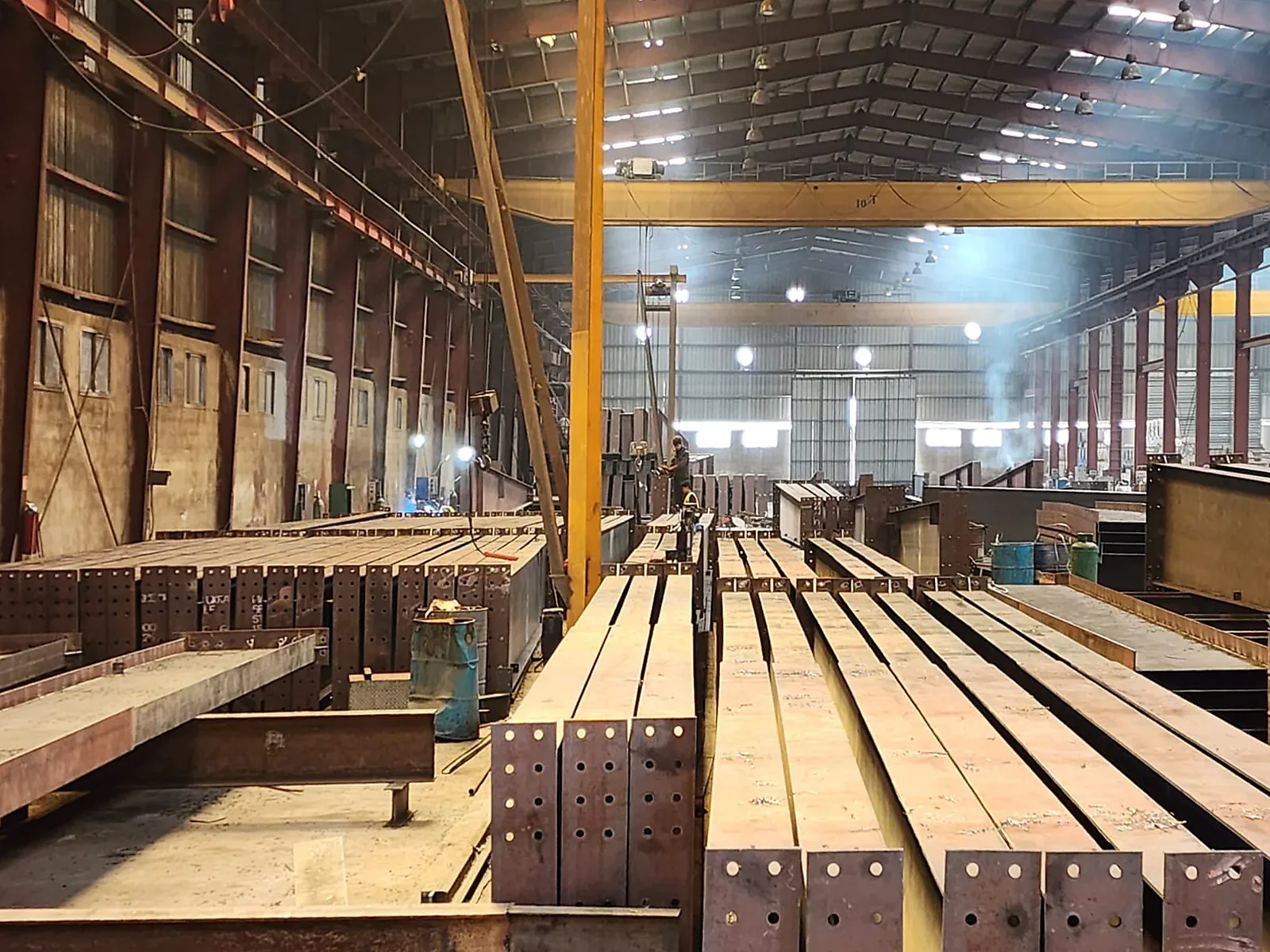 Fabricated steel columns and rafters stacked in the shop with bolted end plates, under a ten-ton bridge crane