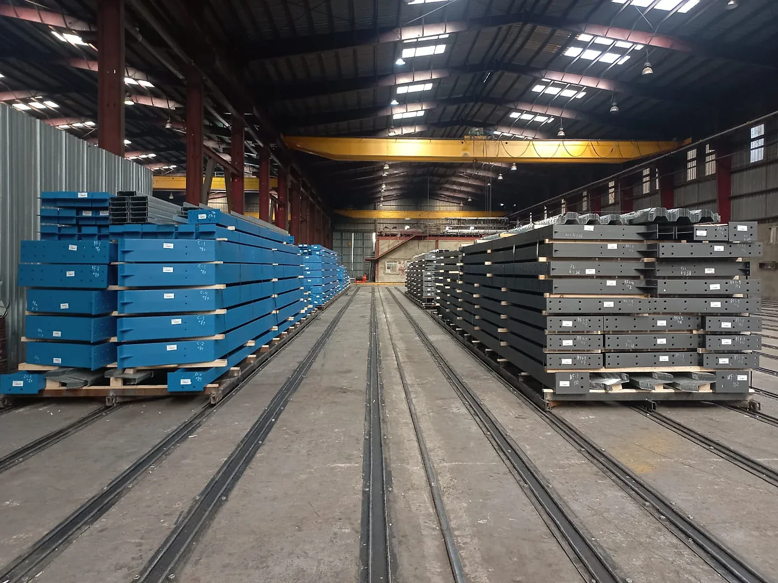 Palletized fabricated steel members with piece-mark labels staged in the plant beneath yellow bridge cranes